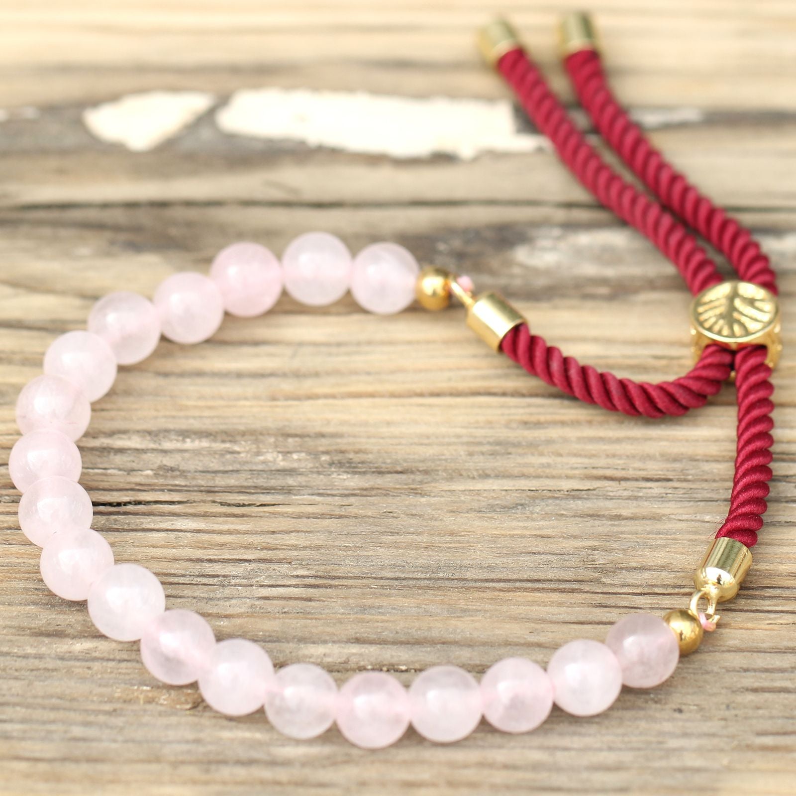 Pulsera de hilo burdeos con piedra de cuarzo rosa en primer plano sobre mesa de madera, accesorio espiritual para armonía emocional.